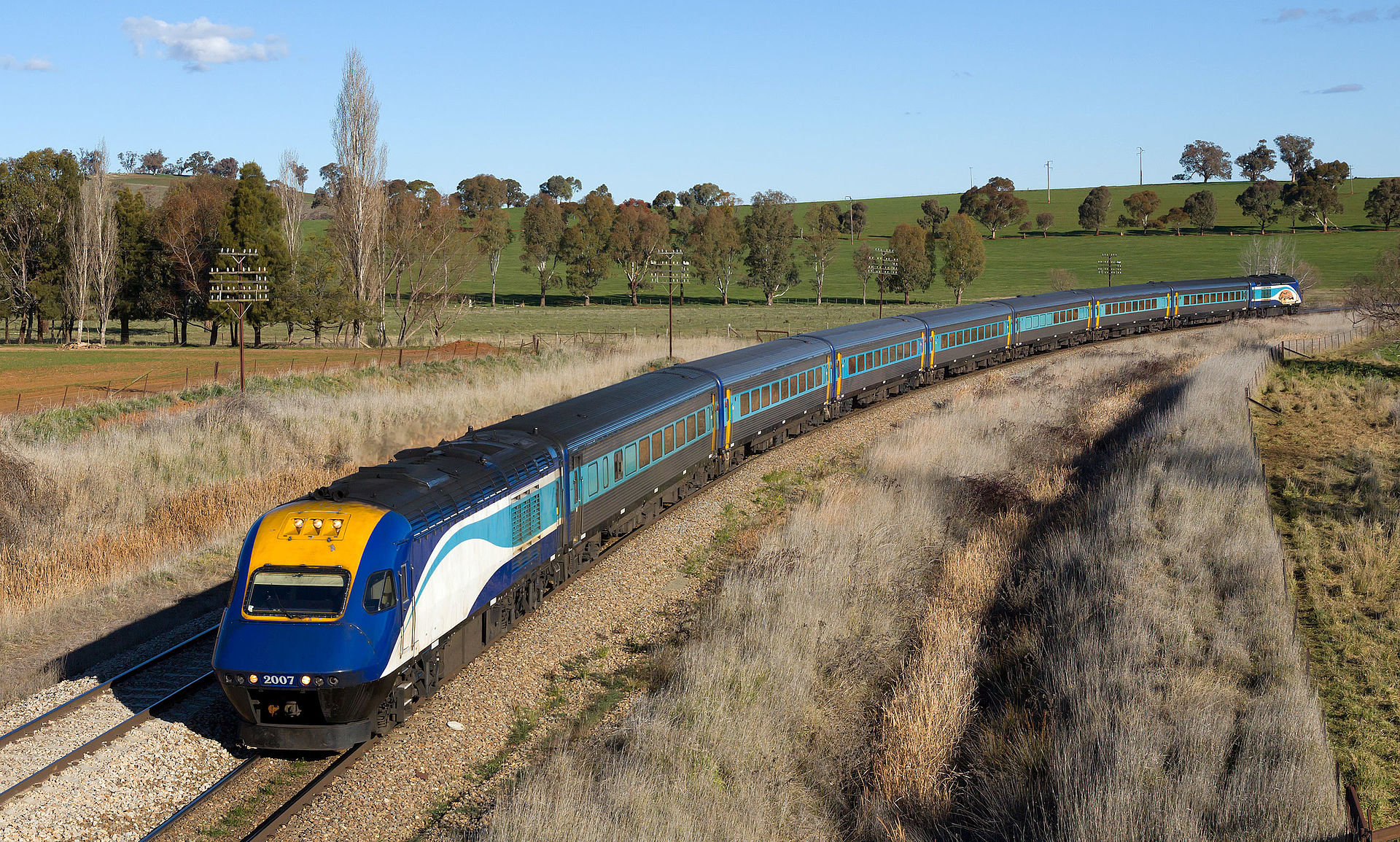 You can travel by train to Wagga Wagga through the great Australian landscape. Photo of blue and yellow train traversing countryside with a background of farmland, trees and bluesky